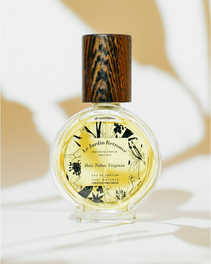 A round bottle of "Le Jardin Retrouvé" Bois Tabac Virginia Limited Edition Eau de Parfum with a wooden cap stands on a white surface against a brown and white abstract background.
