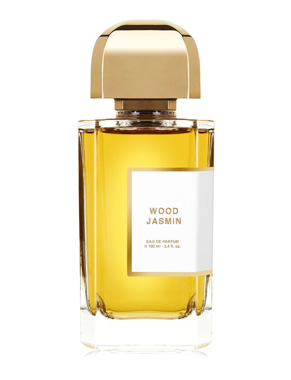 A clear glass bottle of Wood Jasmin Eau de Parfum by BDK Parfums with a gold cap, containing a golden liquid. The white label shows the perfume name and volume details. This precious elixir combines the essence of jasmine with the allure of mysterious woods.