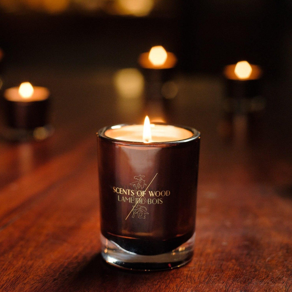 A lit scented candle in a dark glass holder labeled "Scents of Wood" sits on a wooden surface, with the rich aroma of Ebony in Oak wafting in the air, and smaller lit candles blurred in the background.