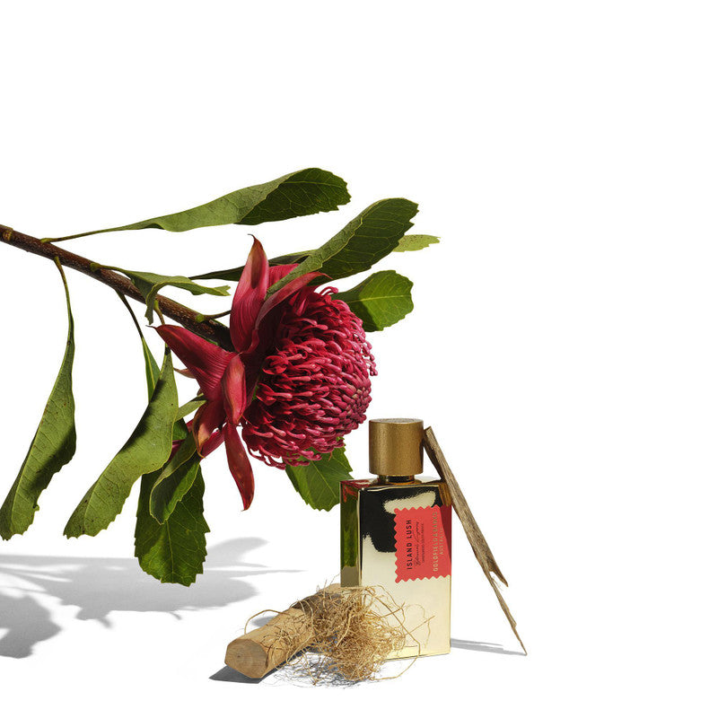 A red flower with green leaves, a small gold bottle of Goldfield & Banks' Island Lush with a red label exuding an exotic fragrance, and a bundle of dried plant material are arranged on a white surface.