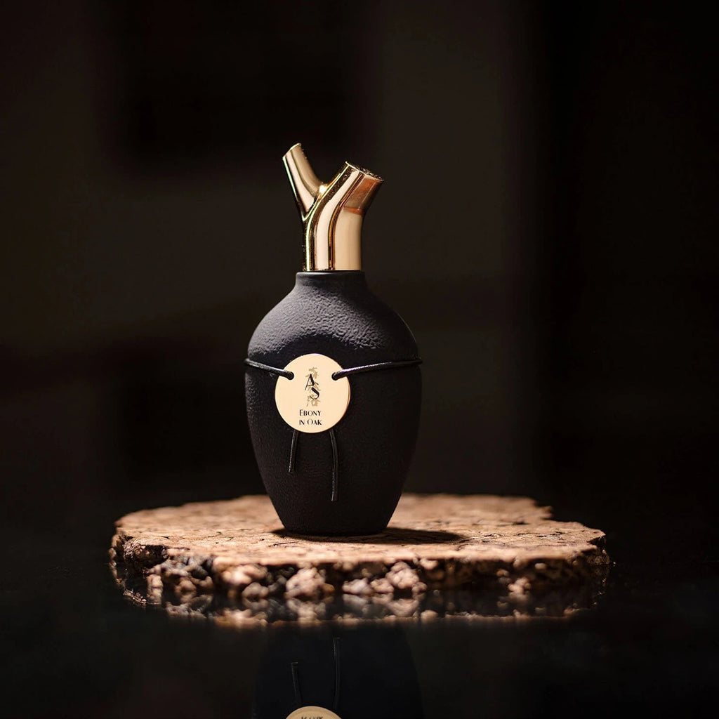 A decorative black bottle of Ebony in Oak by Scents of Wood, with two gold spouts, adorned with a gold emblem featuring text and an image, is set against a dark background on a textured wooden base of ebony wood.
