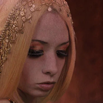 A person with long blonde hair, wearing an ornate golden headpiece and dramatic eye makeup, looks downwards against a dark background, as if stepping out of Ikiryo's Blush of Dawn.