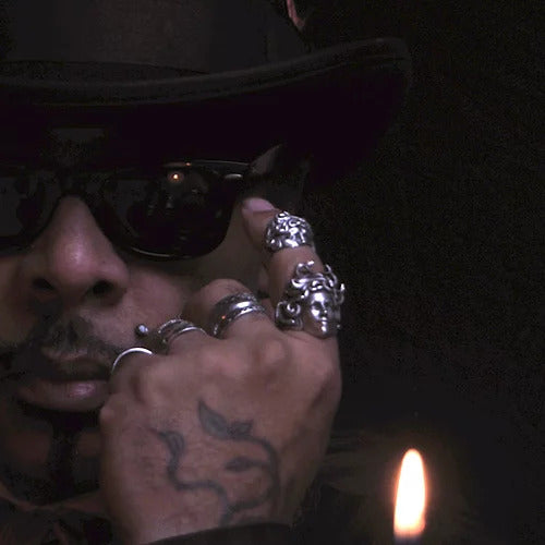 A person wearing dark sunglasses, a hat, and several ornate rings on their fingers is shown in dim lighting with a candle flame in the foreground, exuding an air of intrigue like the Ikiryo CandyMan.