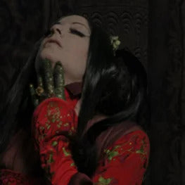 A woman with long black hair, dressed in an ornate red outfit, raises her head with her hand adorned in gold jewelry and green adornments, evoking the rich allure of an Ikiryo Blood Cherry Cordial against a dark, intricate background.