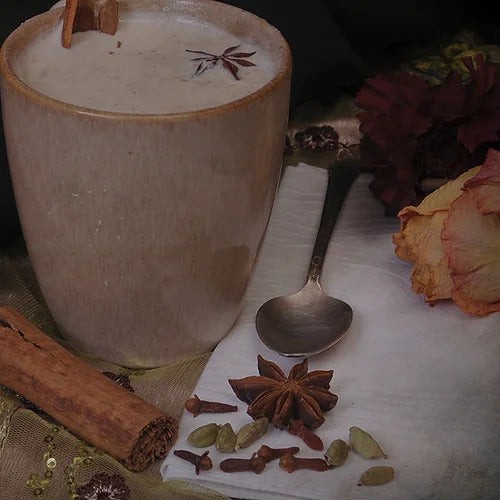 A ceramic mug of spiced latte, reminiscent of fragrant masala chai, with visible star anise and a cinnamon stick, sits next to a spoon on a napkin adorned with cloves and cardamom pods. Nearby, dried flowers add a touch of Crestfallen by Ikiryo.
