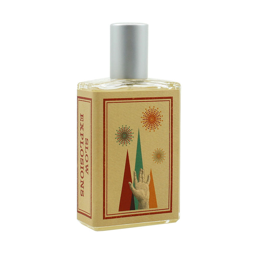 A rectangular bottle of perfume labeled "Slow Explosions" by Imaginary Authors, featuring a design with an outstretched hand and colorful, abstract explosions, enriched with notes of Rose Absolute.