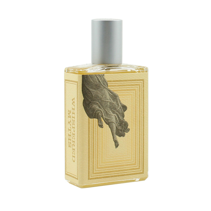 A rectangular perfume bottle with a silver cap and a beige label. The label features an image of a person's arm and hand reaching downward. Text on the label reads "Whispered Myths" by Imaginary Authors. Infused with notes of Cedarwood, this fragrance captures an essence both timeless and pure.