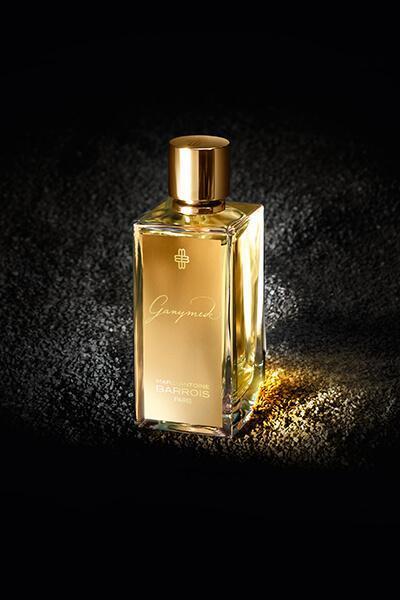 A rectangular perfume bottle with a gold cap and label stands against a dark, textured background. The label reads "Ganymede, Marc-Antoine Barrois.