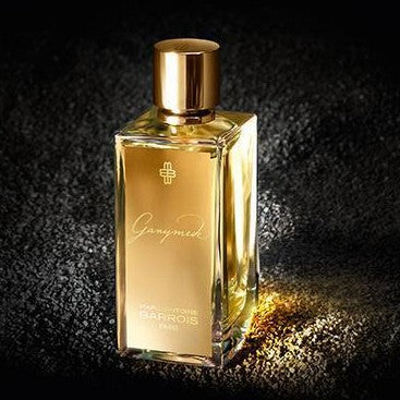 A rectangular perfume bottle with a gold cap and label stands against a dark, textured background. The label reads "Ganymede, Marc-Antoine Barrois.