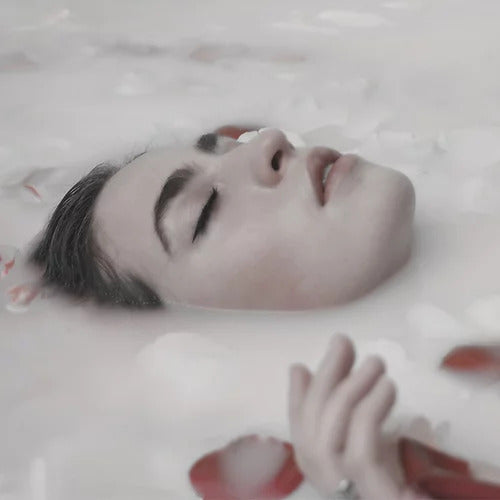 A person with closed eyes is partially submerged in milky water with scattered rose petals, emanating a serene aura reminiscent of AGDLENAWRIKM (Dreamwalking) by Ikiryo.