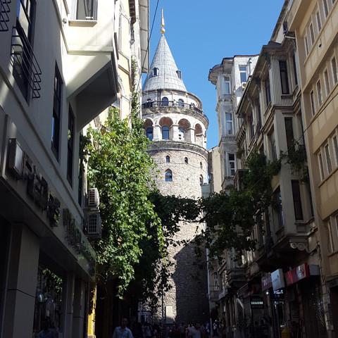 A narrow street lined with buildings leads to the historic Galata Tower, its conical roof standing proudly in Istanbul's ancient cityscape. Under a clear blue sky, the air carries a hint of bergamot reminiscent of Gallivant Perfumes' "Istanbul by Gallivant.
