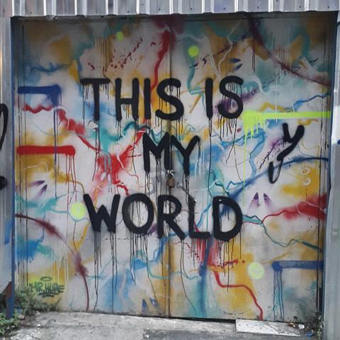 A vibrant mural on a metal wall, emblazoned with the bold black phrase "This is my world," showcases an array of abstract shapes and drips in red, blue, yellow, and various other hues. The scene captures the spirit of Istanbul by Gallivant from Gallivant Perfumes, evoking the essence of an ancient city infused with the tangy scent of bergamot.