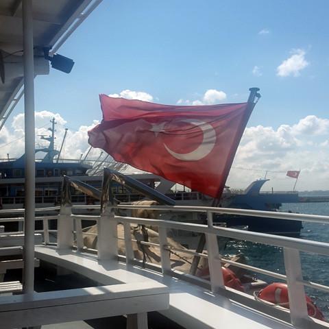 A Turkish flag waves on a ferry docked by the water with other boats visible in the background under a partly cloudy sky, reminiscent of the ancient city evoked by Gallivant Perfumes' "Istanbul," where scents of bergamot once filled the air.