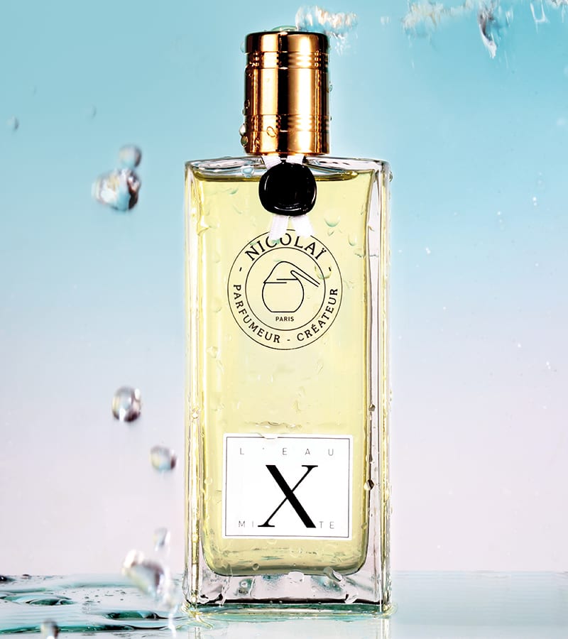 A bottle of Nicolaï L'Eau Mixte perfume with a clear liquid and gold cap is surrounded by water droplets, set against a blue background. The scent features refreshing notes of Sicilian lemon, adding a citrus twist to its allure.