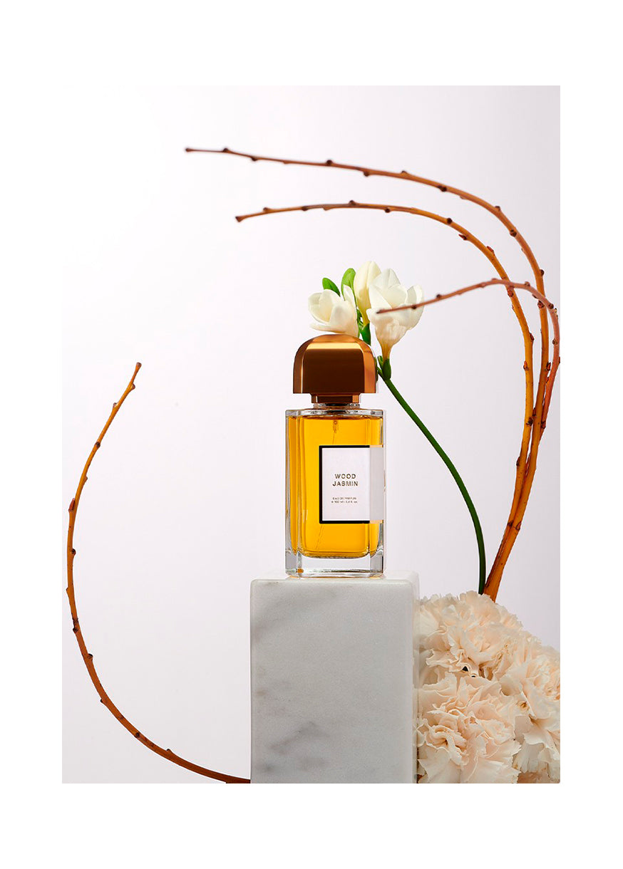 A perfume bottle with a golden cap and amber liquid stands on a white pedestal. Delicate Jasmine flowers and curved brown branches are arranged around it, creating an elegant display that whispers of mysterious woods.