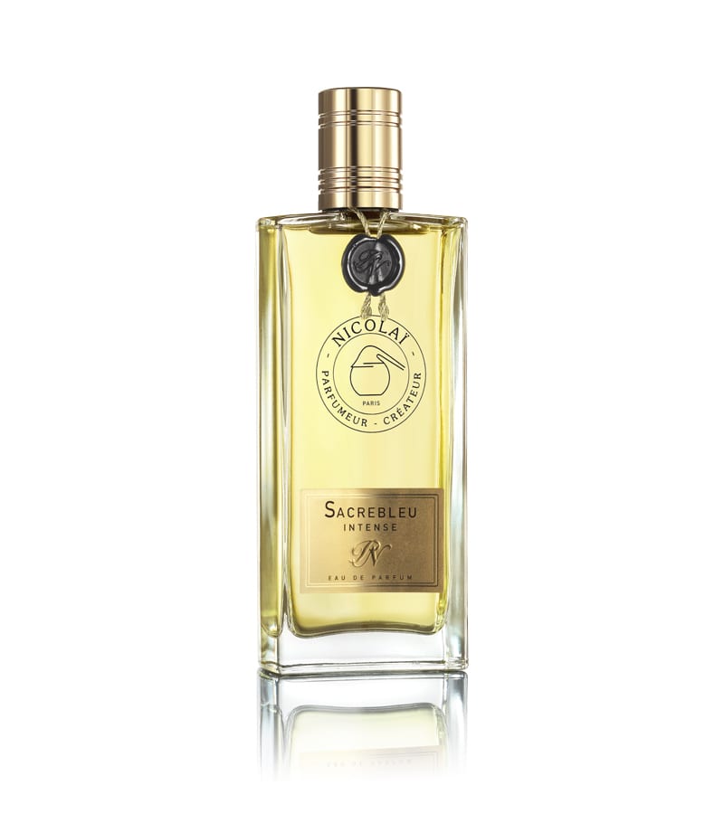 A rectangular glass bottle of Nicolaï's Sacrebleu Intense perfume with a gold cap and label, containing a golden liquid. The bottle features a round medallion on the neck, hinting at its enchanting powdery floral fragrance with notes of jasmine and white flowers.
