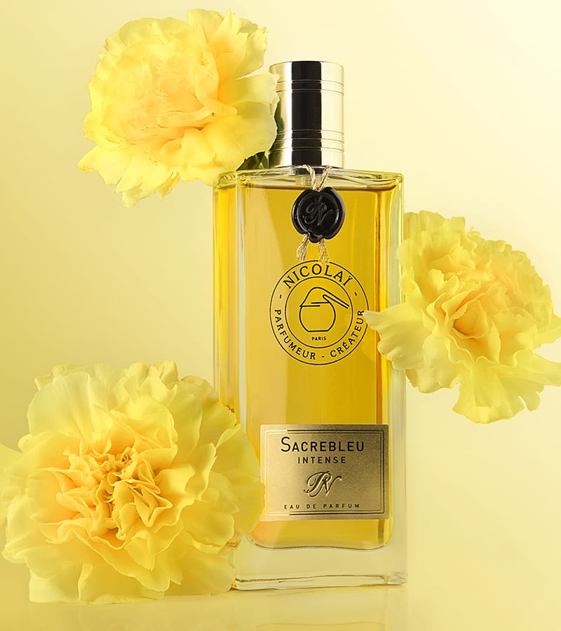 A bottle of "Sacrebleu Intense" by Nicolaï, elegantly encircled by yellow and white flowers, is showcased against a vivid yellow background. This powdery floral fragrance emanates a harmonious blend of jasmine that heightens its allure.
