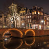 Nighttime view of a lit bridge over a canal with surrounding illuminated buildings and bare trees in a city setting, the air subtly tinged with the allure of Amsterdam by Gallivant from Gallivant Perfumes.