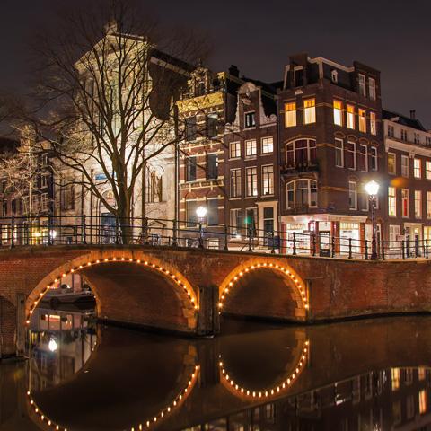 Nighttime view of a lit bridge over a canal with surrounding illuminated buildings and bare trees in a city setting, the air subtly tinged with the allure of Amsterdam by Gallivant from Gallivant Perfumes.