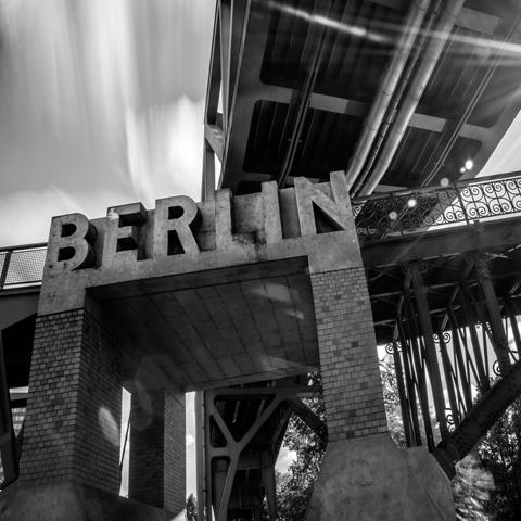 A black and white photo captures a large concrete sign reading "Berlin" mounted under a metal bridge structure, evoking the bold essence of Berlin by Gallivant from Gallivant Perfumes.