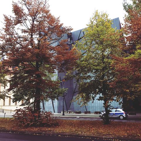 A modern building with a metallic facade behind two autumn-colored trees, with a police car parked on the street in front. The scene is reminiscent of Berlin by Gallivant from Gallivant Perfumes, capturing the essence of grapefruit, clementine, and lemon notes filling the crisp air.