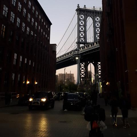 View of Manhattan Bridge framed by two brick buildings in Brooklyn at dusk, with a few people and a car visible on the street below, giving off the refreshing summer scent of Brooklyn by Gallivant from Gallivant Perfumes.