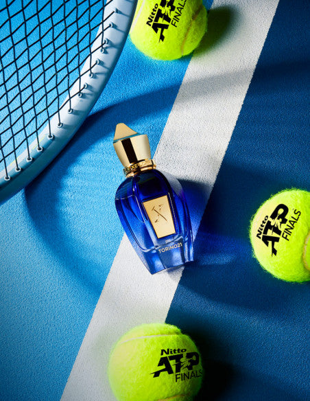 A blue Xerjoff Torino 21 perfume bottle is placed on a blue and white tennis court, surrounded by three yellow tennis balls branded with "Nitto ATP Tennis Finals," and part of a tennis racket is visible in the top left corner.