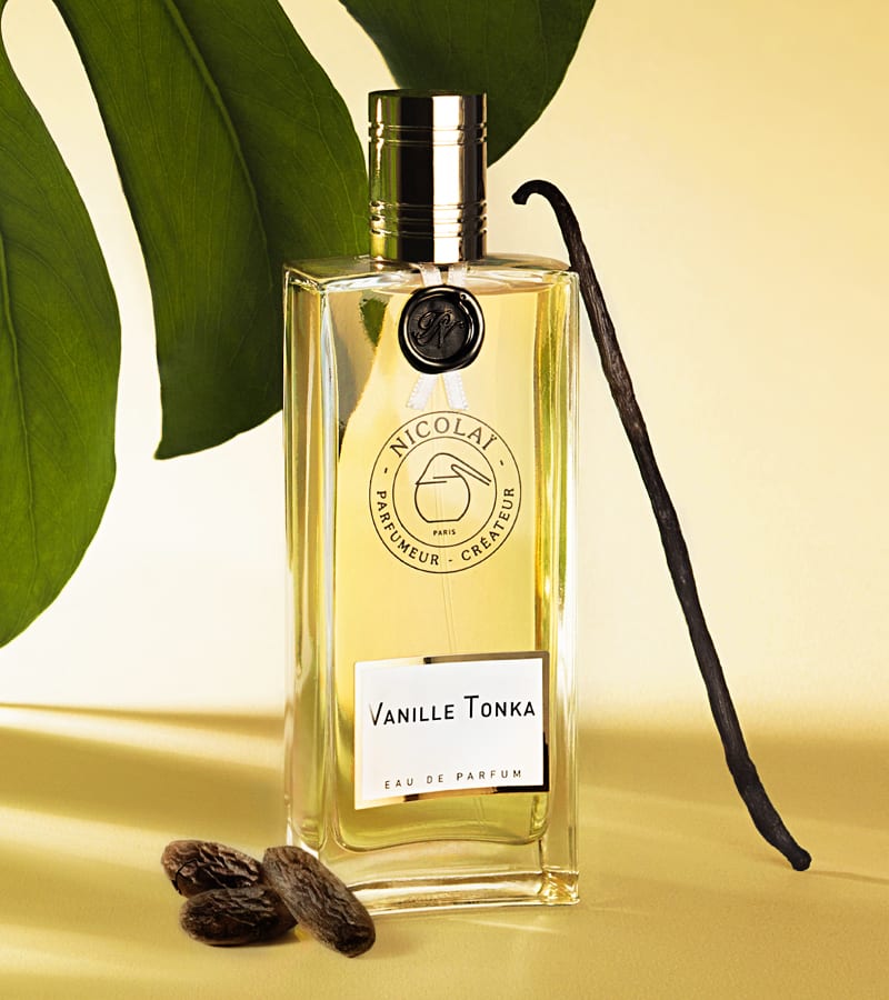 A bottle of Vanille Tonka by Nicolaï is placed against a yellow background with a large leaf, a vanilla bean, and some tonka beans beside it. Subtle notes of incense waft in the air, adding to the fragrance's allure.