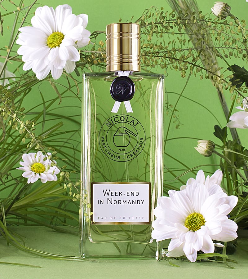 A bottle of "Week-End In Normandy" eau de toilette by Nicolaï is surrounded by white daisies, green foliage, and the fresh scent of cut grass against a vibrant green background.