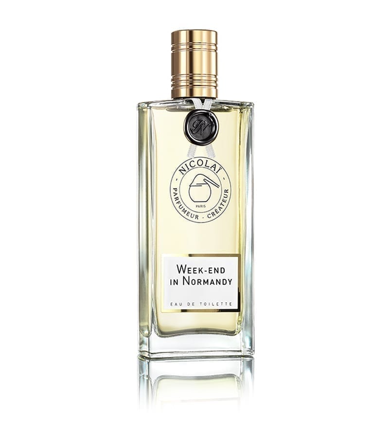 A clear glass bottle of "Week-End in Normandy" Eau de Toilette by Nicolaï with a gold and black cap, evoking the fresh scent of cut grass and lily of the valley.